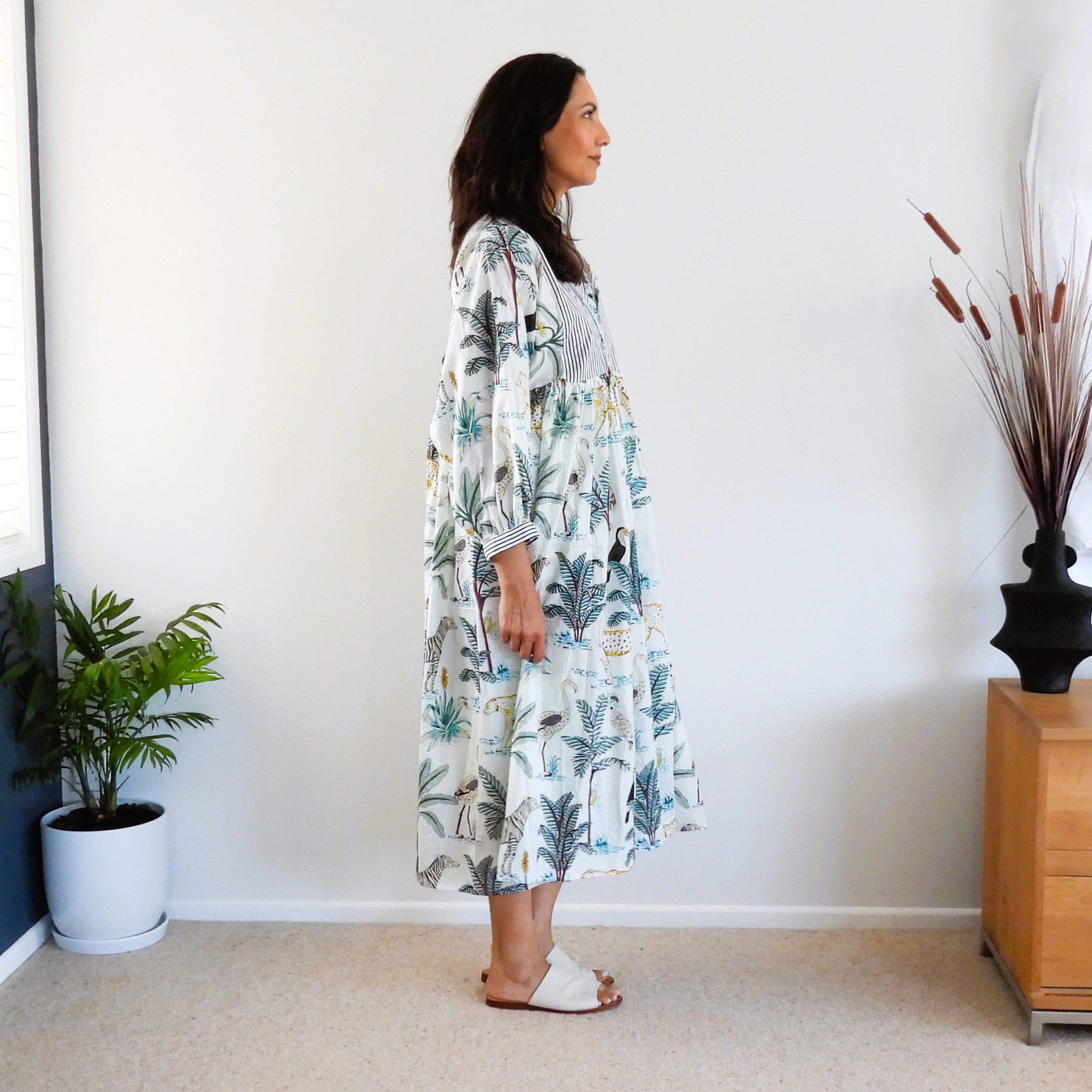 A woman wearing a long-sleeved, knee-length maxi dress with a tropical palm leaf print in shades of green, blue, and yellow on a white background.