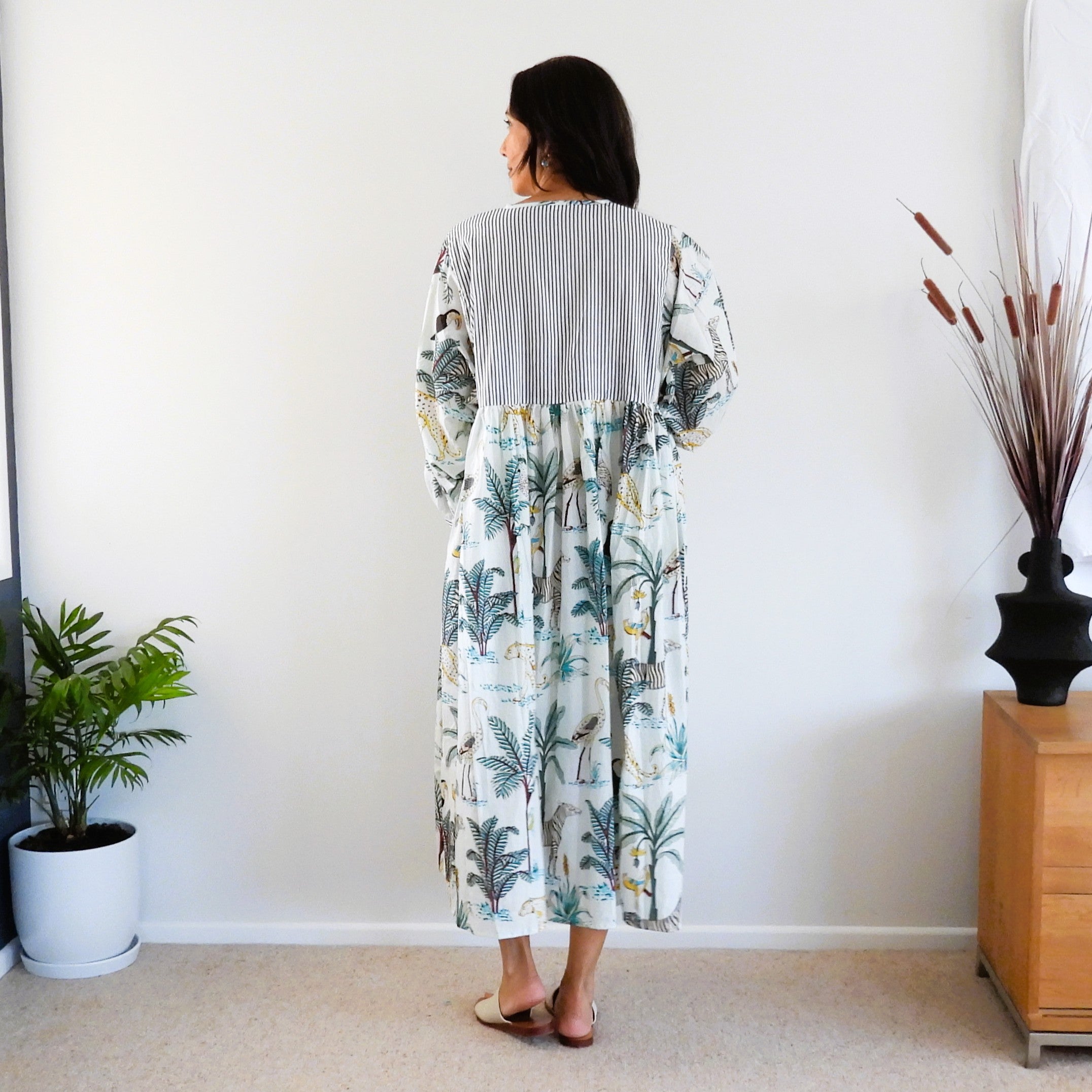 A woman stands with her back to the camera wearing a long-sleeved midi dress with a gray striped yoke and a tropical palm tree print skirt. 