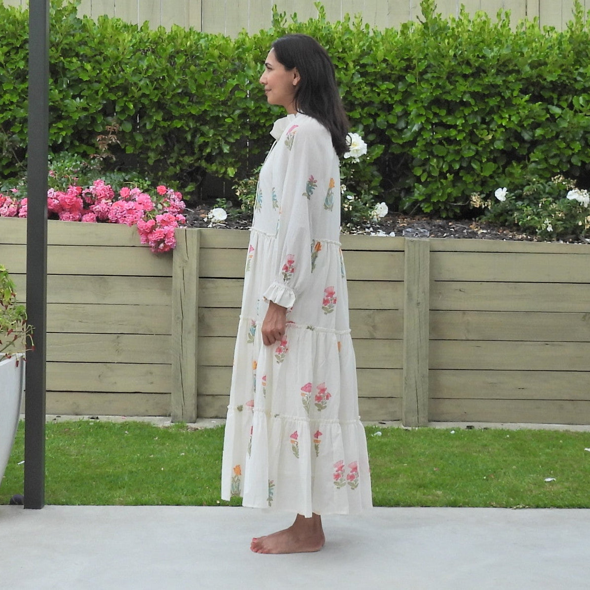 Woman in a white dress standing under a patio cover with a garden and fence in the background