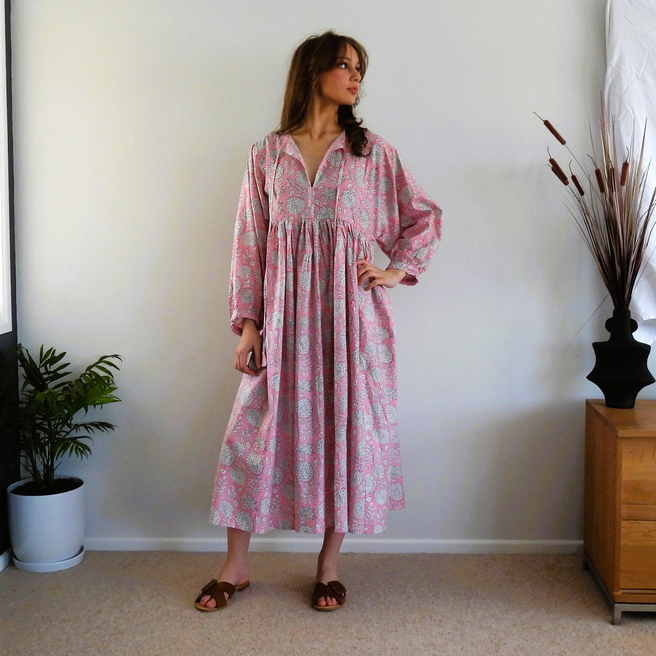A woman stands in a room wearing a pink and white paisley midi dress with long sleeves and a gathered waist. She is wearing brown slide sandals, with a potted plant on the left and a wooden cabinet with dried grass on the right.