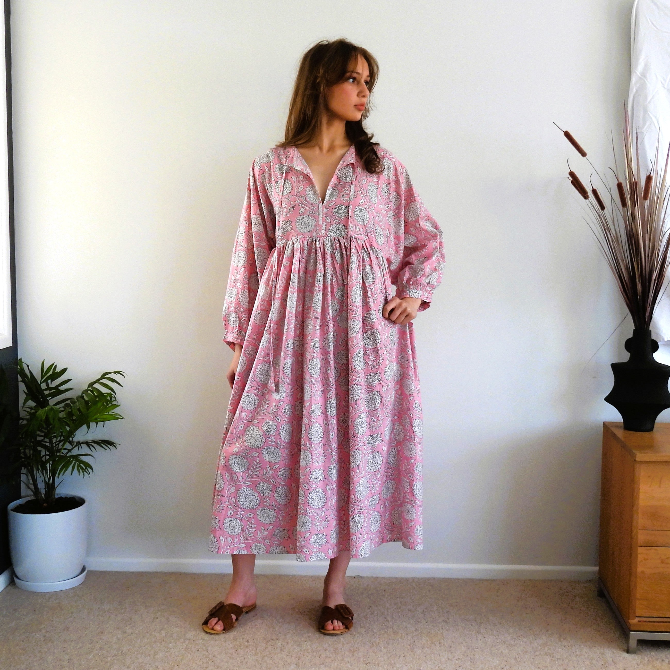 A woman wearing a pink long-sleeve midi dress with a light blue and white paisley pattern, standing in a room with a white wall, beige carpet, a potted plant, and a wooden side table.