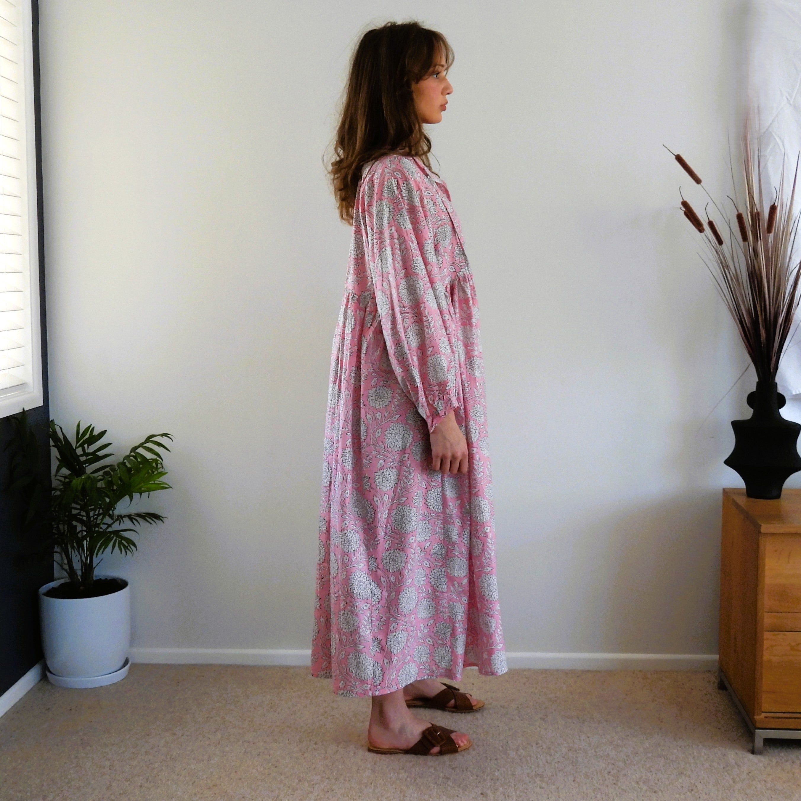 A woman stands in profile wearing a long-sleeve, midi-length pink dress with a light green and white paisley pattern in a room with beige carpet, a white wall, a potted plant, and a wooden side table.