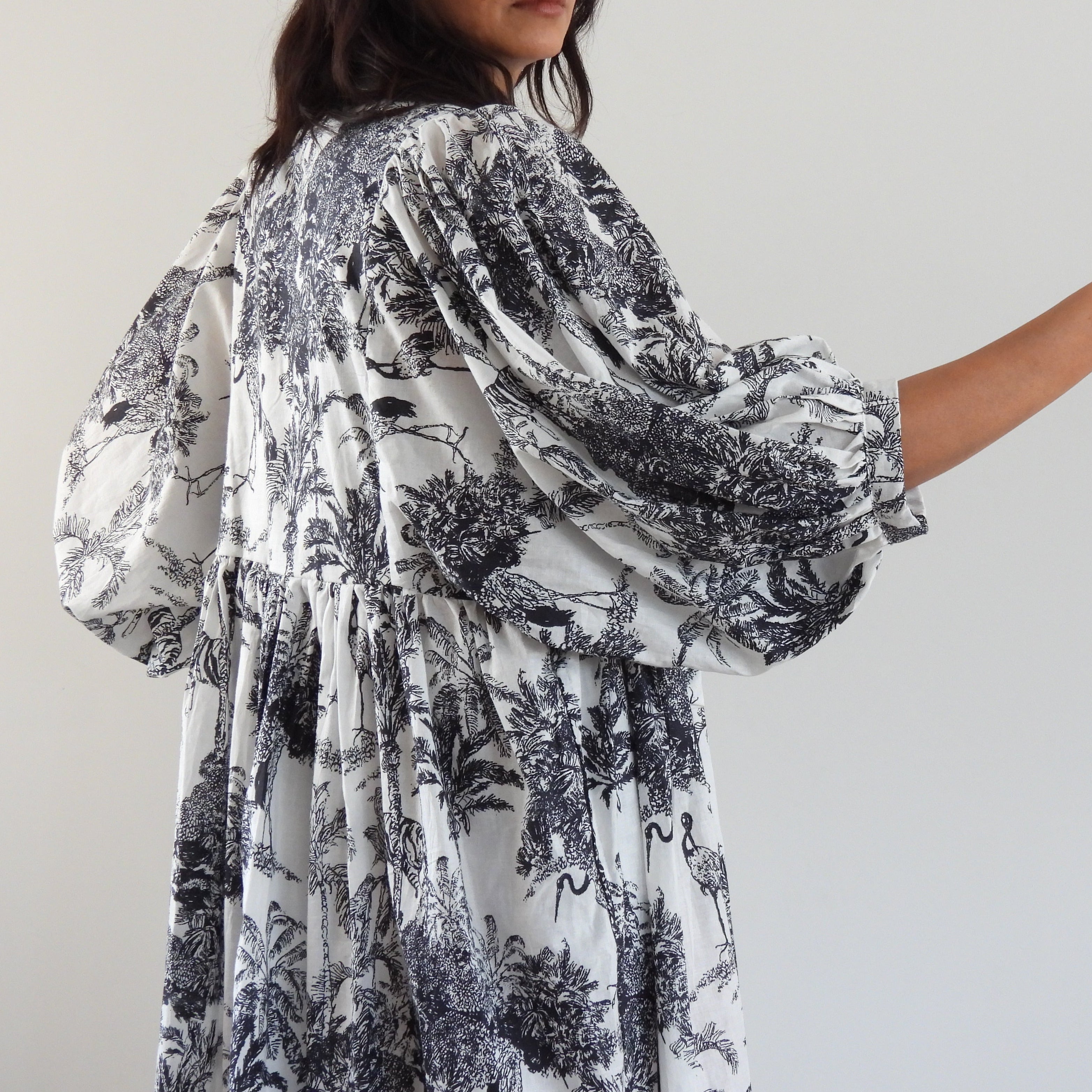 A woman wearing a white long-sleeve dress with a black toile-style botanical print, shown from the back and side against a plain light gray wall.