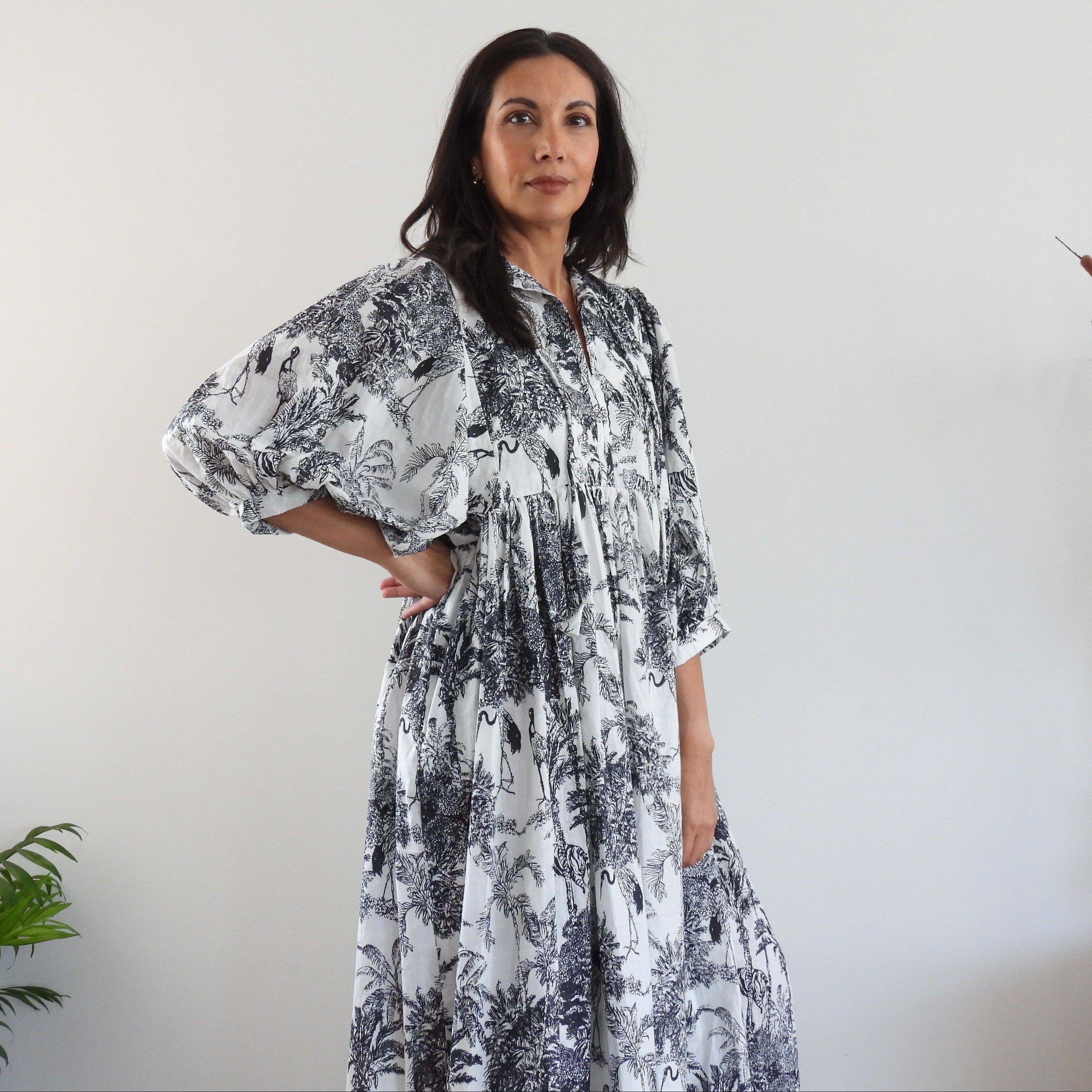 A woman modeling a long-sleeved, loose-fitting maxi dress with a black and white toile-style print, standing in a minimalist room with a white wall, a plant, and a decorative vase.