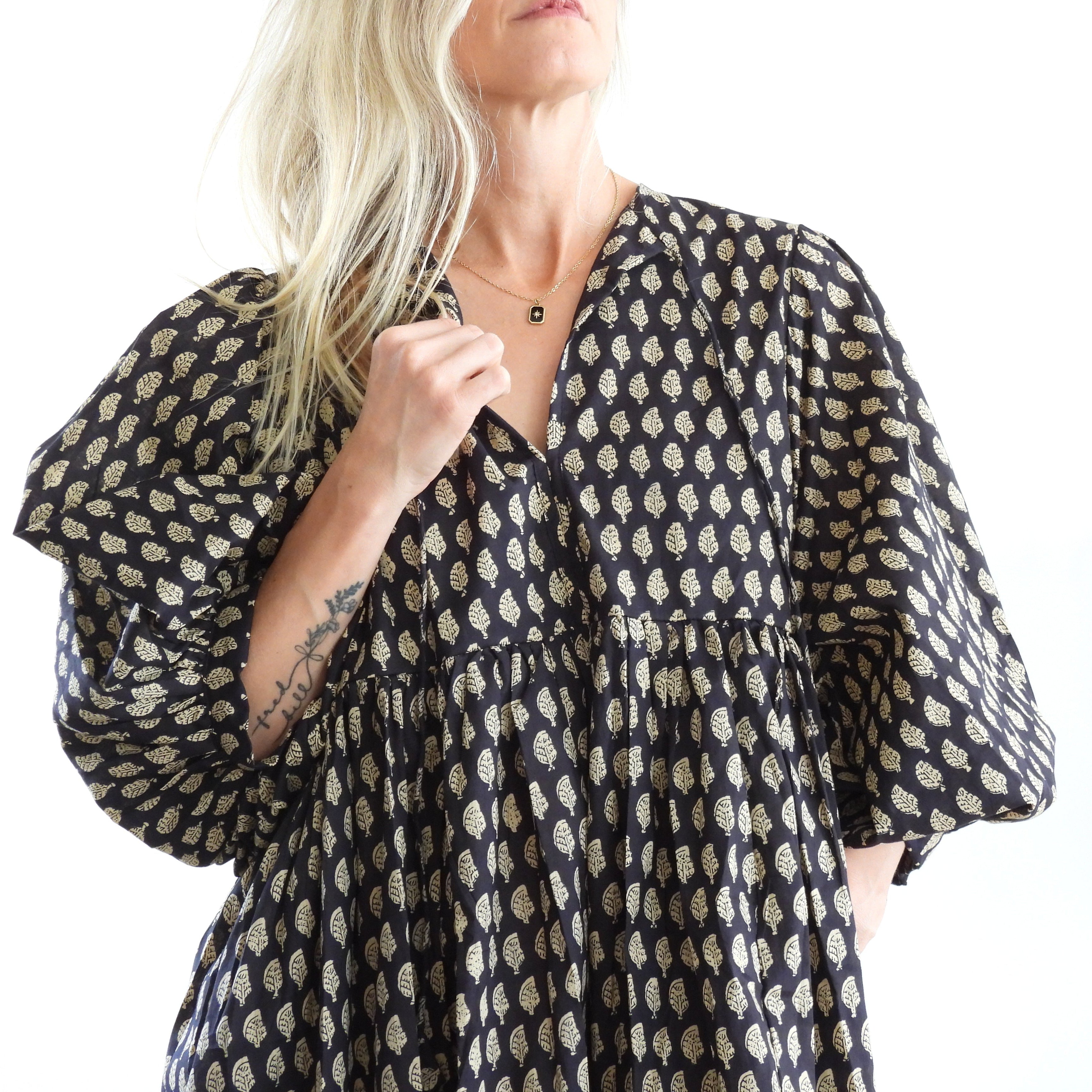 A woman wearing a loose black dress with a beige leaf pattern, featuring voluminous elbow-length sleeves and a tiered skirt, set against a bright white background.