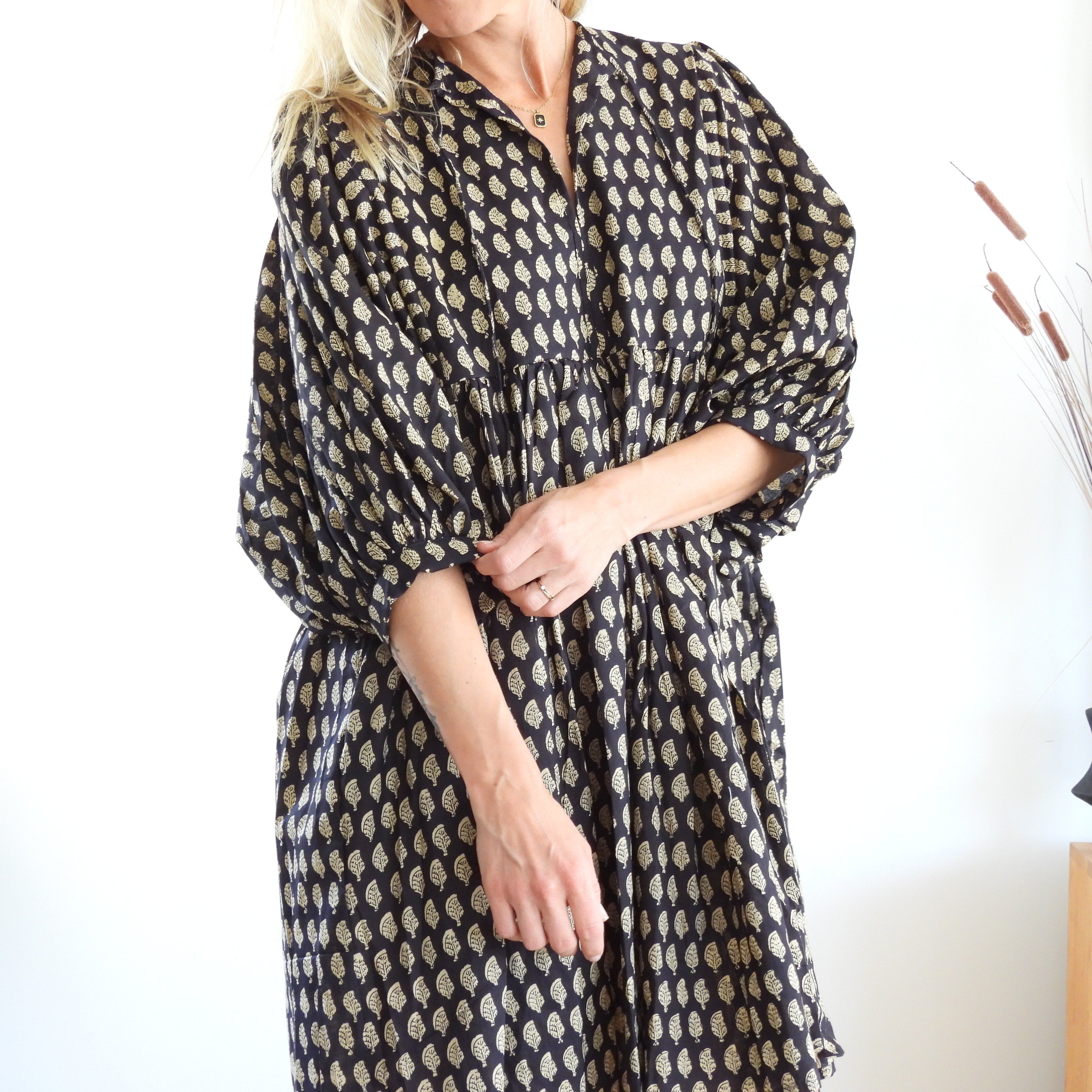 A woman wearing a loose-fitting black dress with a beige geometric pattern, standing in a bright indoor setting with a white wall, a wooden side table, and a black vase with dried grass.