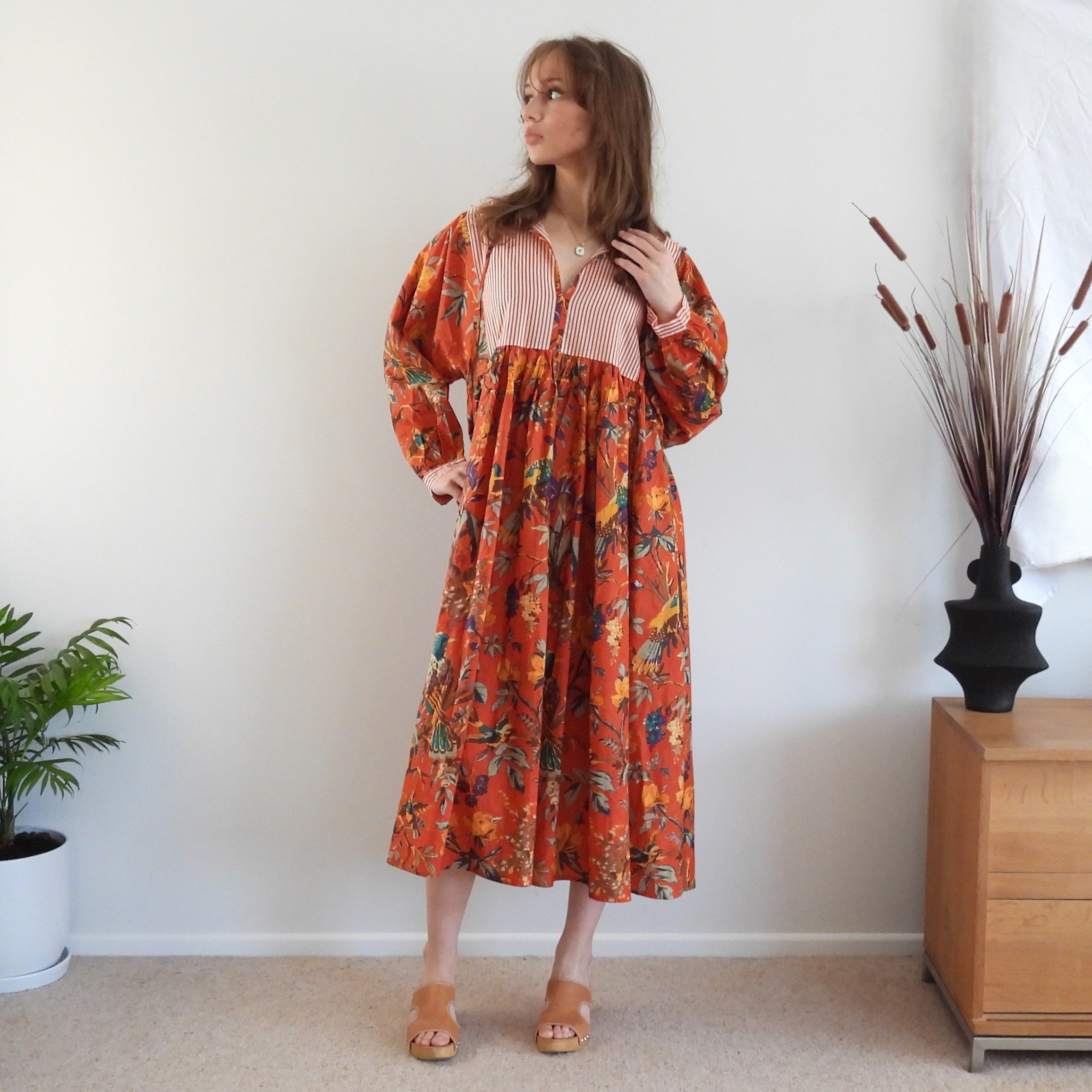 A woman stands in a room wearing a long-sleeved midi dress with a vibrant orange, red, and yellow floral pattern and a striped yoke. She is wearing tan wedge sandals, standing in front of a white wall with a wooden cabinet and a potted plant on the right.