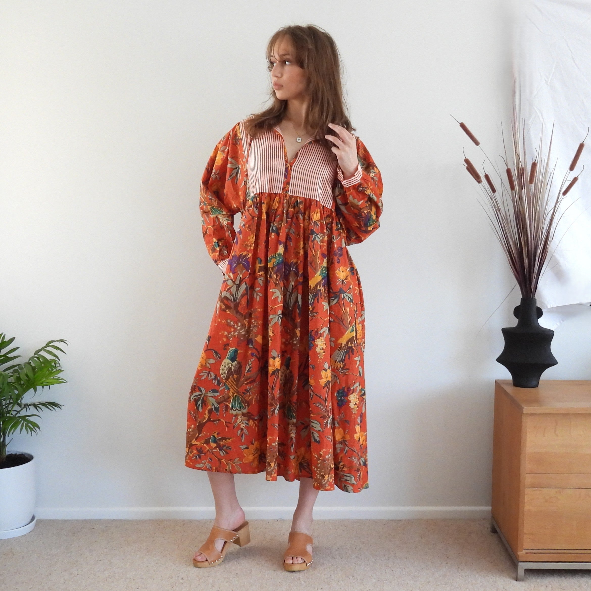 A woman stands indoors wearing a long-sleeve midi dress with a vibrant orange-red base and a multicolor floral print, featuring a pink striped V-neck bodice. She is posed in a room with a white wall, a wooden cabinet, and a potted plant.