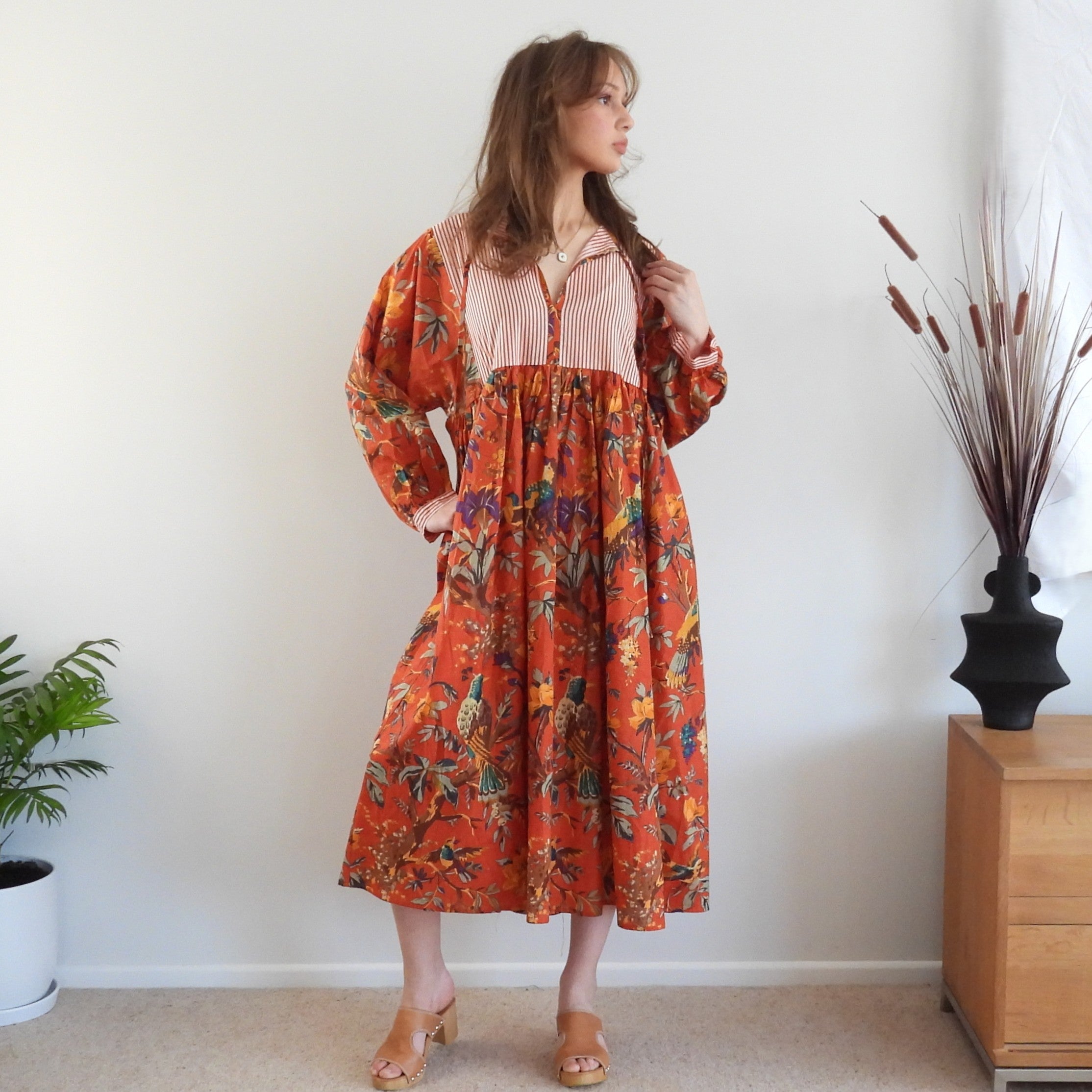 A woman stands indoors wearing a long-sleeve midi dress with a bright orange-red base and an allover multicolor floral and bird print, plus a light pink striped yoke. The room has a white wall, beige carpet, a wooden cabinet, and a black vase with dried grass.