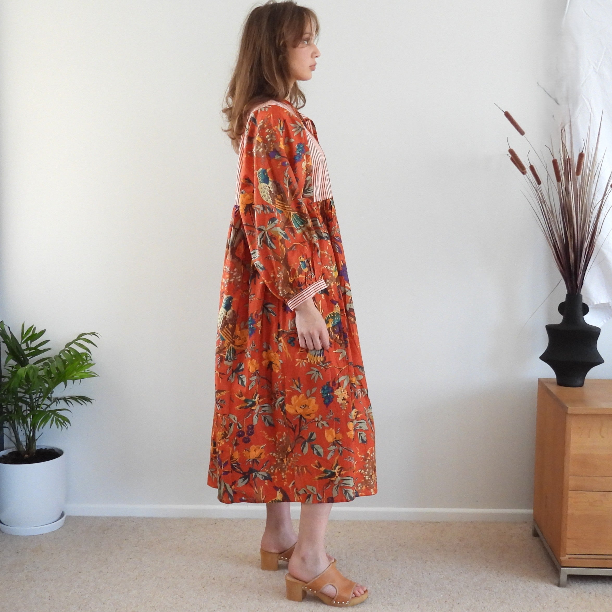 A woman stands in profile wearing a long-sleeved midi dress with a vibrant orange-red floral print, paired with tan block-heel sandals, set against a neutral indoor background with a plant and wooden cabinet.