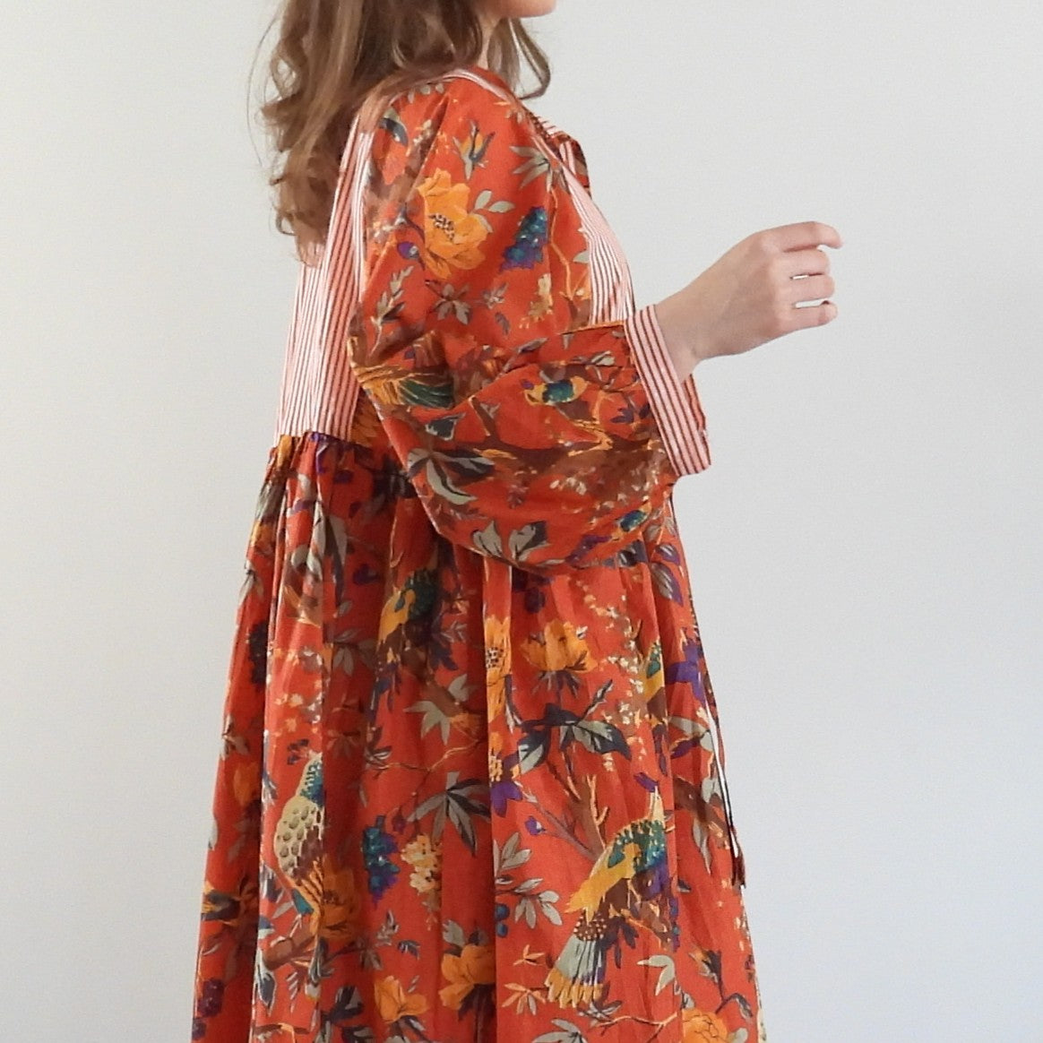 A woman wearing a long-sleeved orange dress with a colorful floral and bird print, featuring a striped bodice and waist tie, shown from a side-back angle against a plain light gray background.