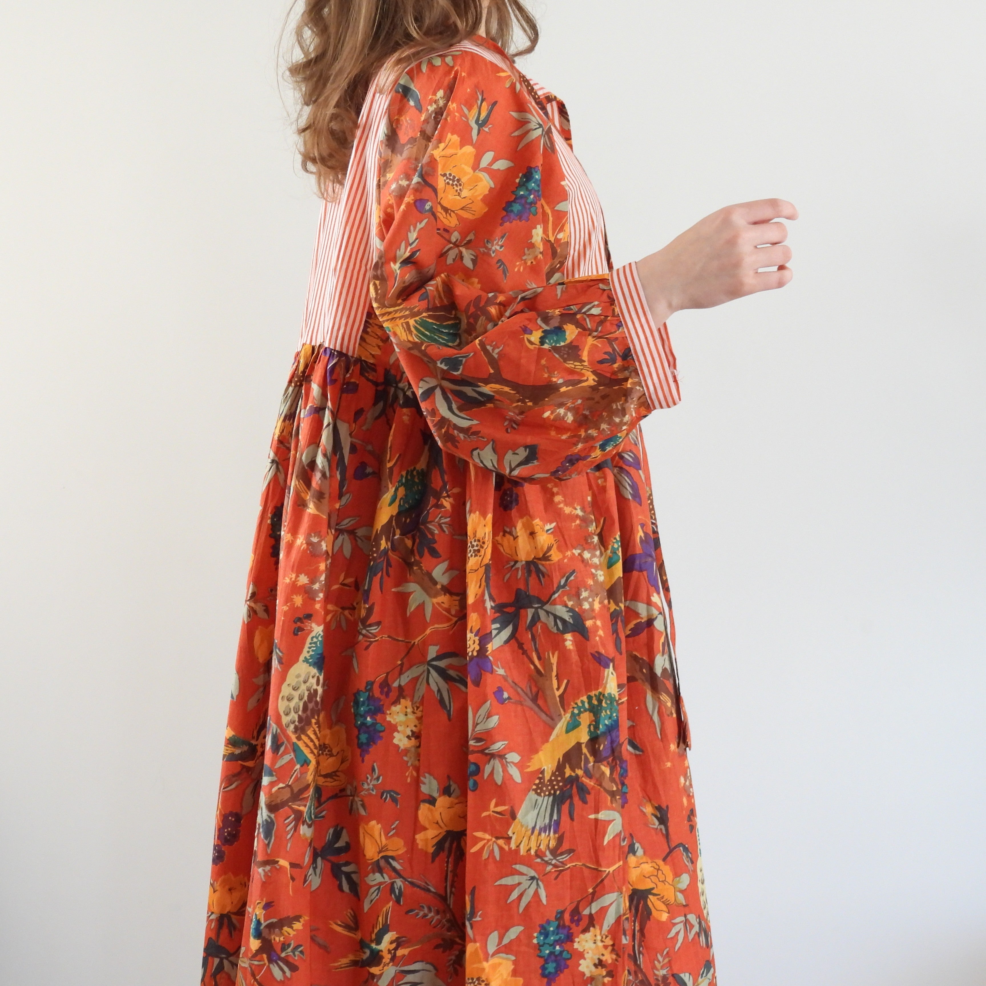 A woman wearing a long-sleeved orange dress with a colorful floral and bird print, featuring a striped bodice and cuffs, photographed against a plain white background.
