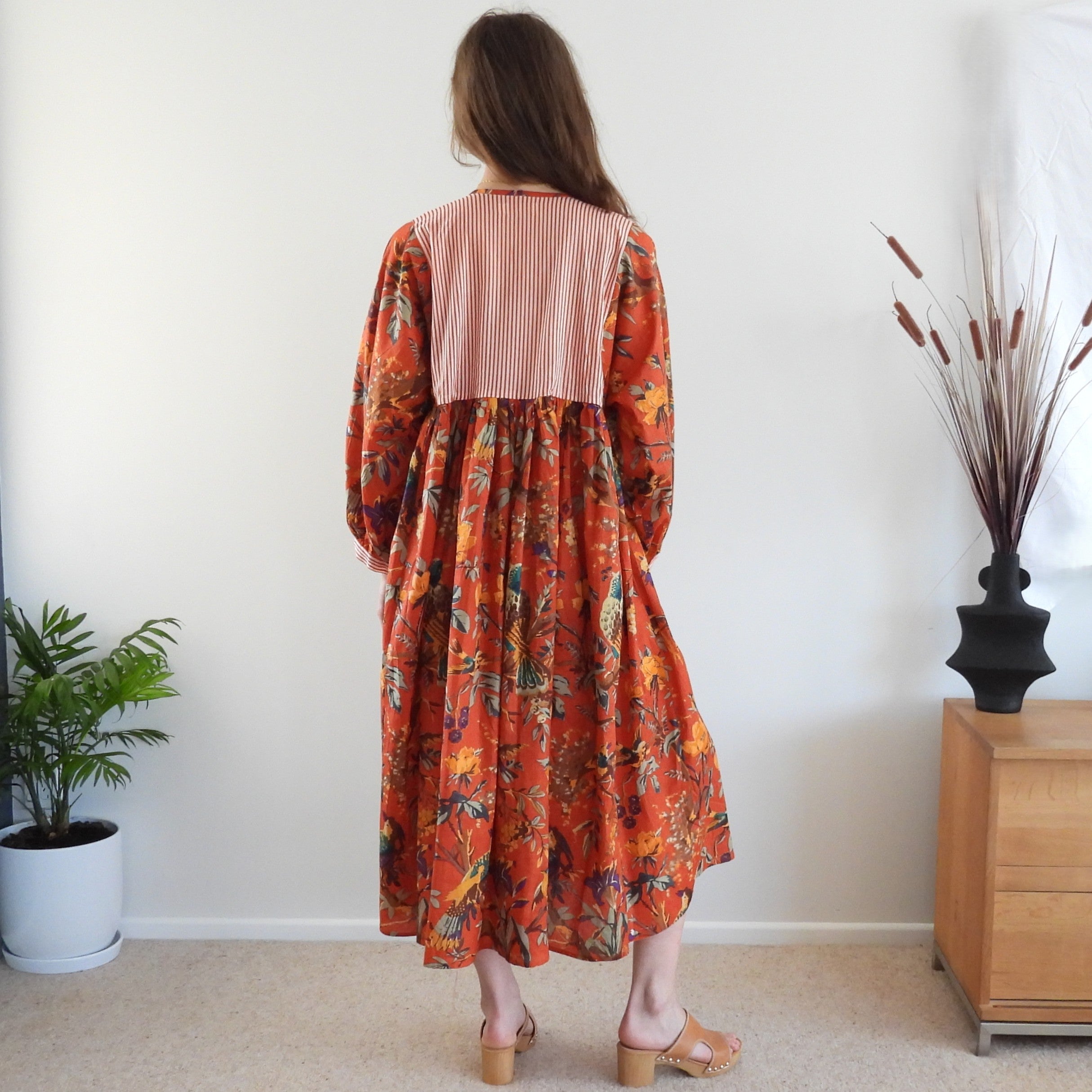 A woman stands with her back to the camera wearing a long-sleeved midi dress with a striped back panel and a vibrant orange floral print skirt. She is in a minimalist room with a white wall, beige carpet, a potted plant, and a wooden side table with dried grass.