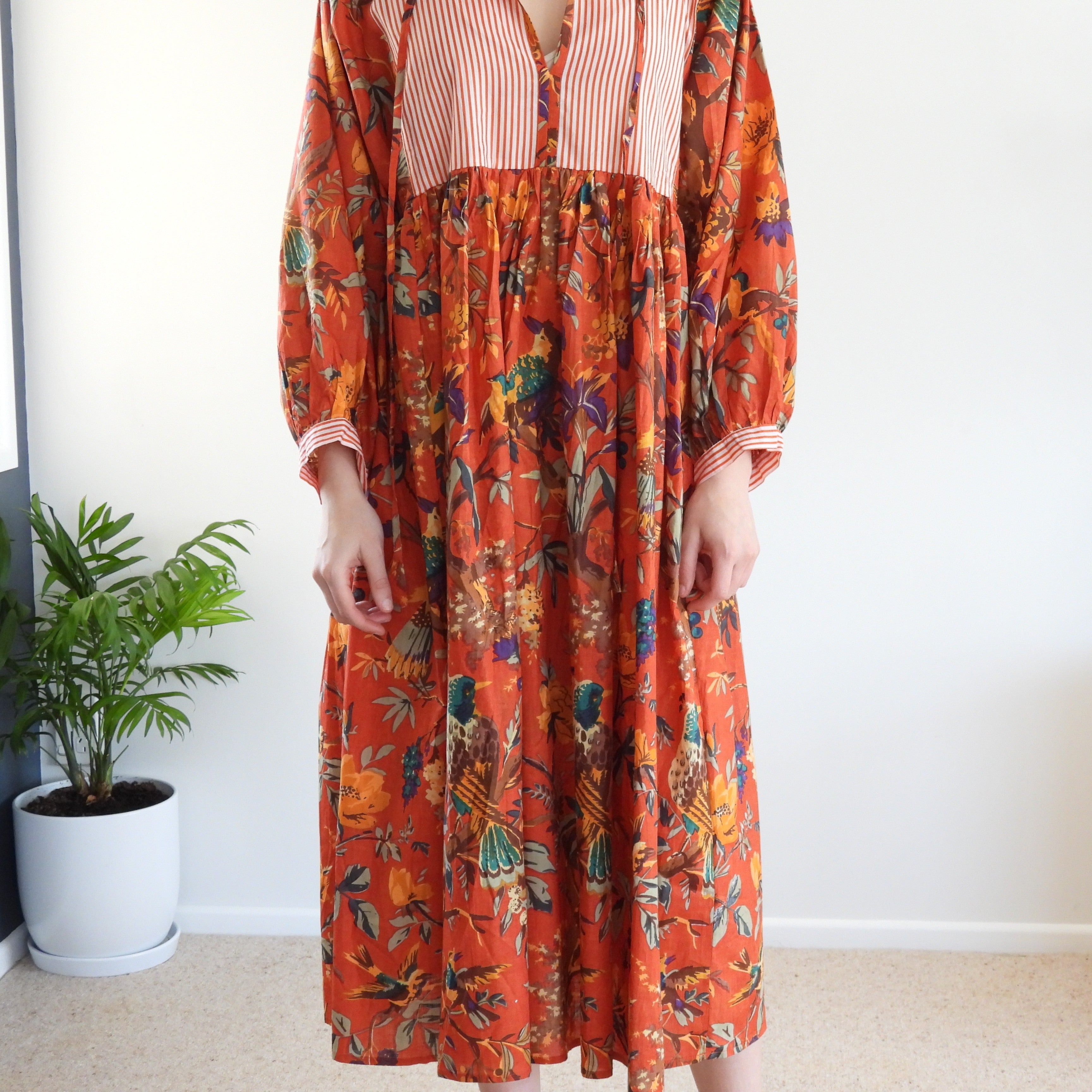 A woman wearing a long-sleeved midi dress with a vibrant orange background and a multicolor tropical print, featuring a striped bodice. She is standing indoors next to a potted plant.