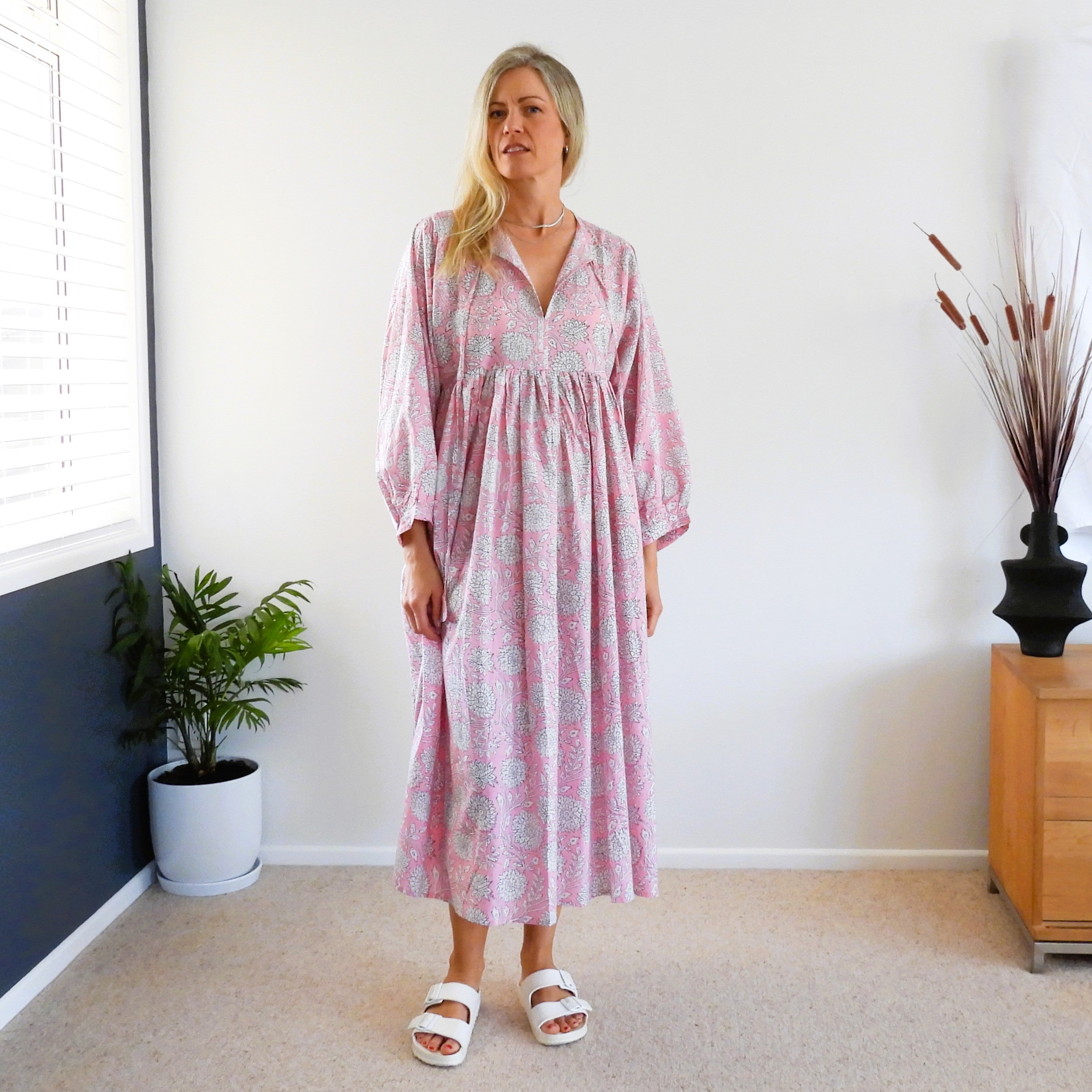 A woman stands in a bright room wearing a pink and white floral midi dress with three-quarter sleeves and white slide sandals.