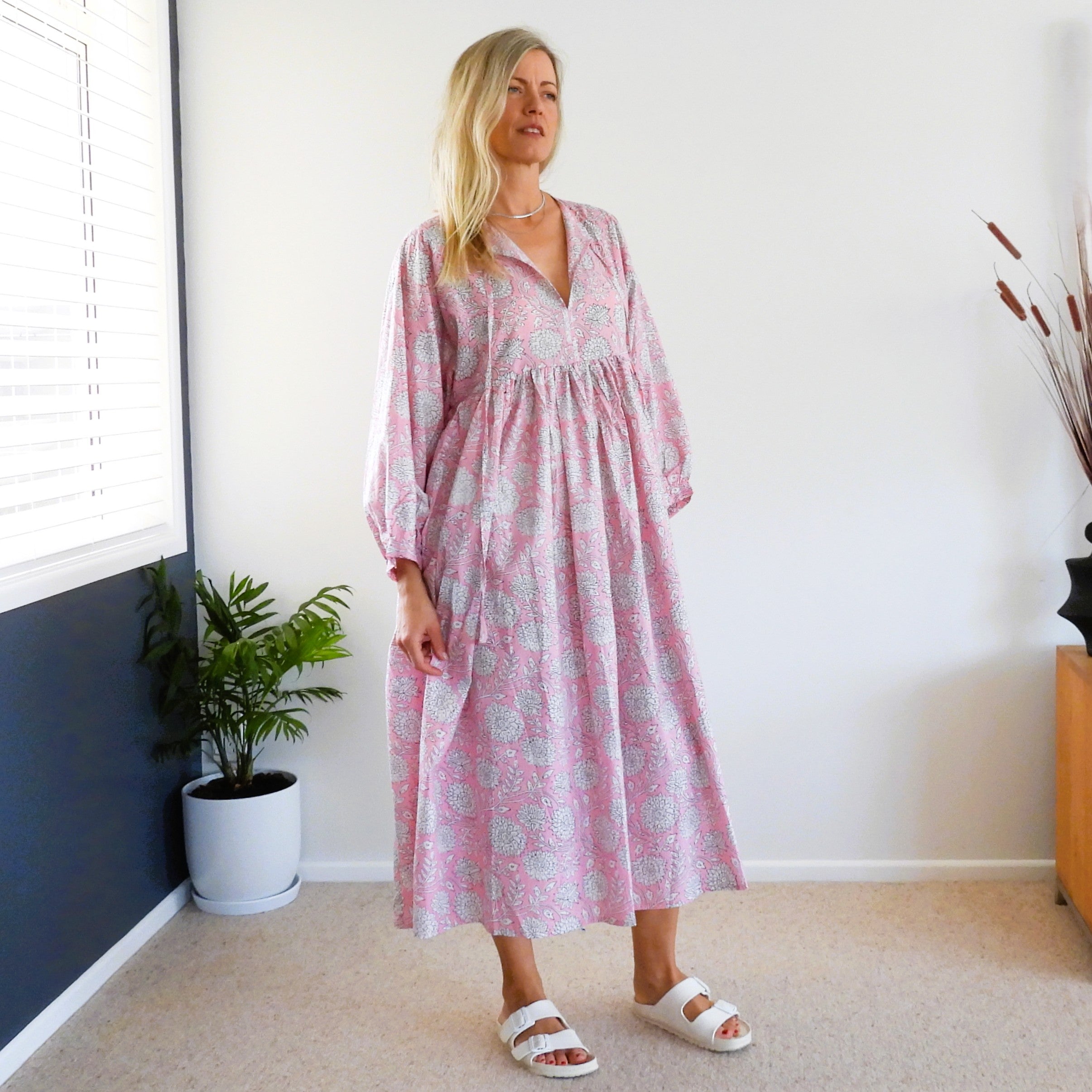 A woman wearing a pink and white floral midi dress with three-quarter sleeves and white slide sandals, standing in a brightly lit room with a window, potted plant, and beige carpet.