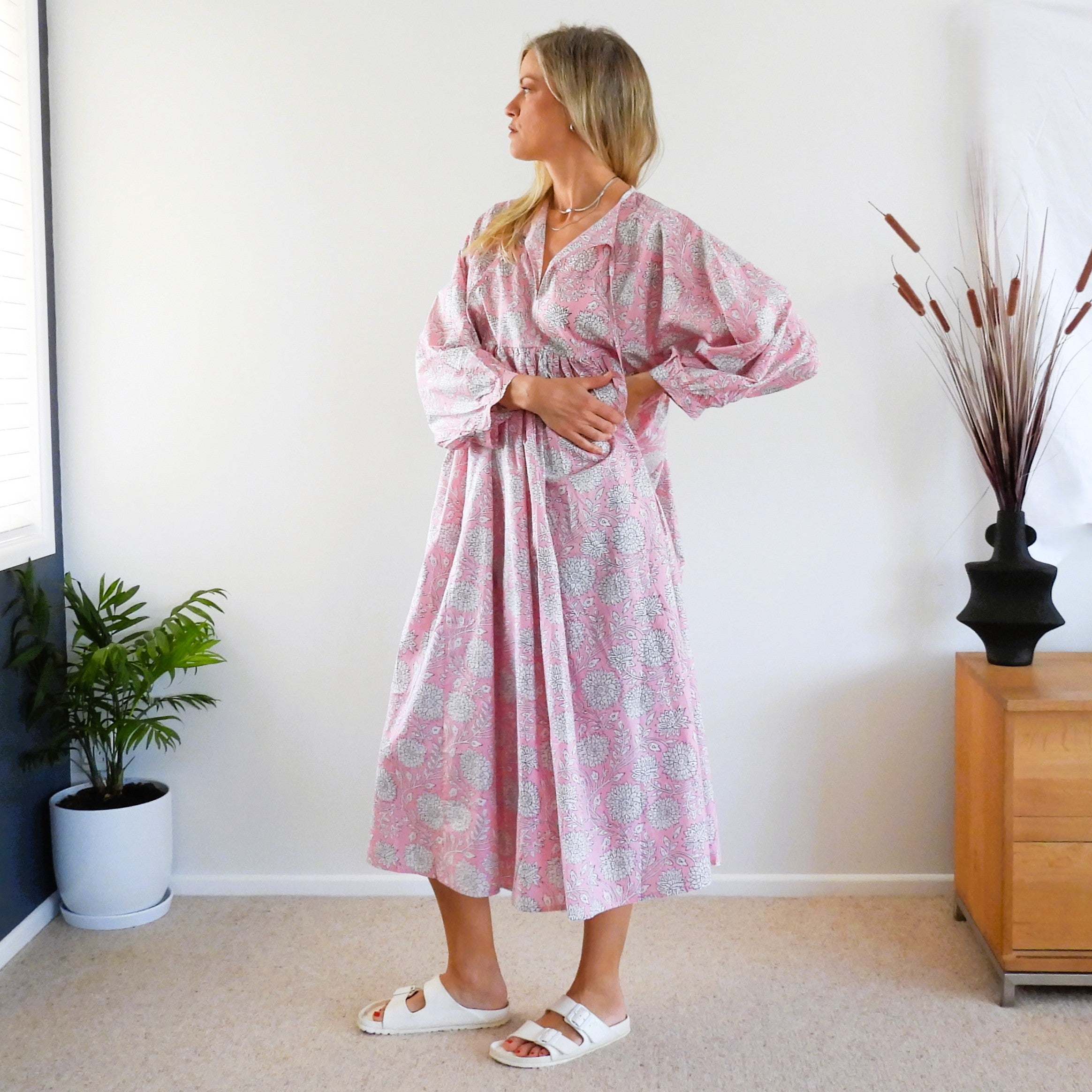A woman wearing a pink and white patterned kaftan dress with voluminous sleeves, standing in a brightly lit room with a white wall, a potted plant, and a wooden cabinet.