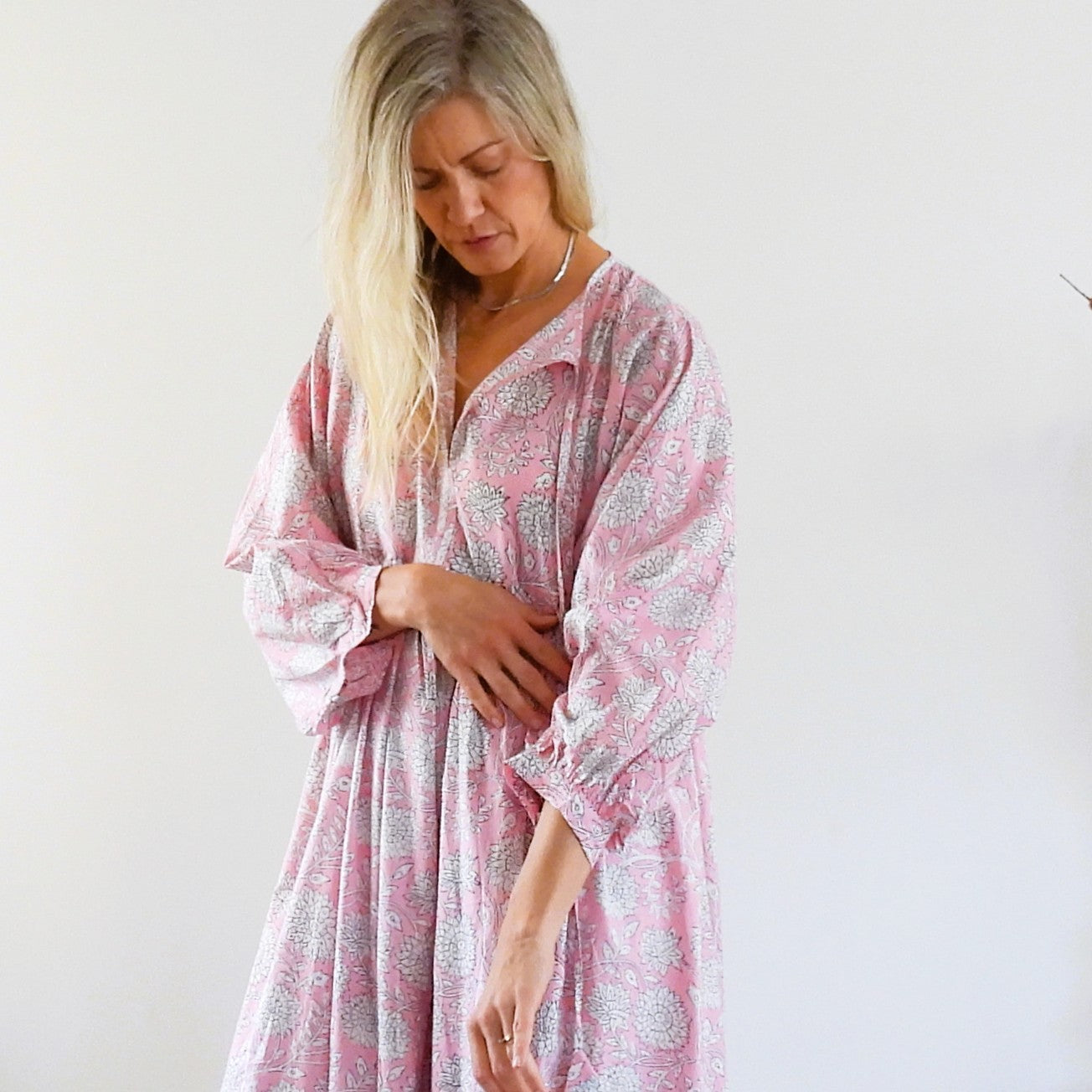 A woman wearing a pink and gray paisley print kaftan dress, standing against a plain white wall.
