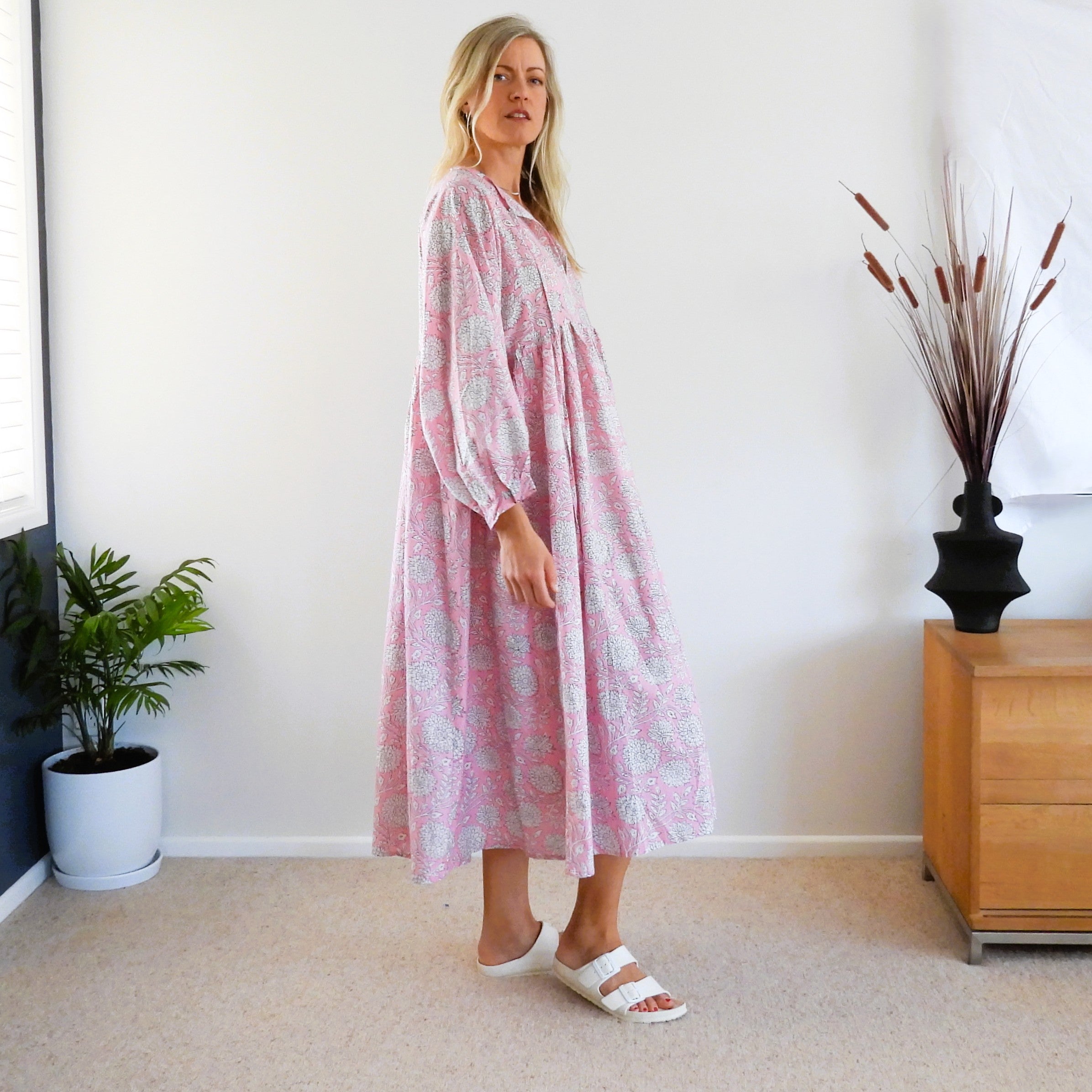 A woman modeling a long-sleeved, midi-length pink dress with a light blue floral pattern, standing in a brightly lit room with a beige carpet, white wall, a potted plant, and a wooden side table.