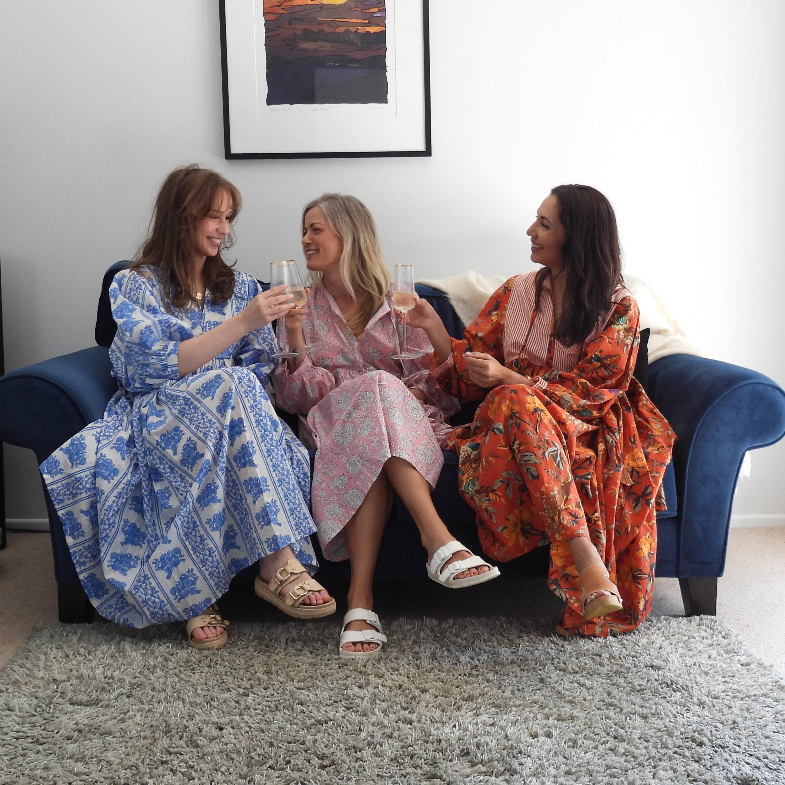 Three women sitting on a blue sofa in a bright living room, wearing long patterned robes or kaftan-style dresses in blue, pink, and orange-red tones. A gray shag rug covers the floor, with a lamp, books, wall art, and a glass door visible.