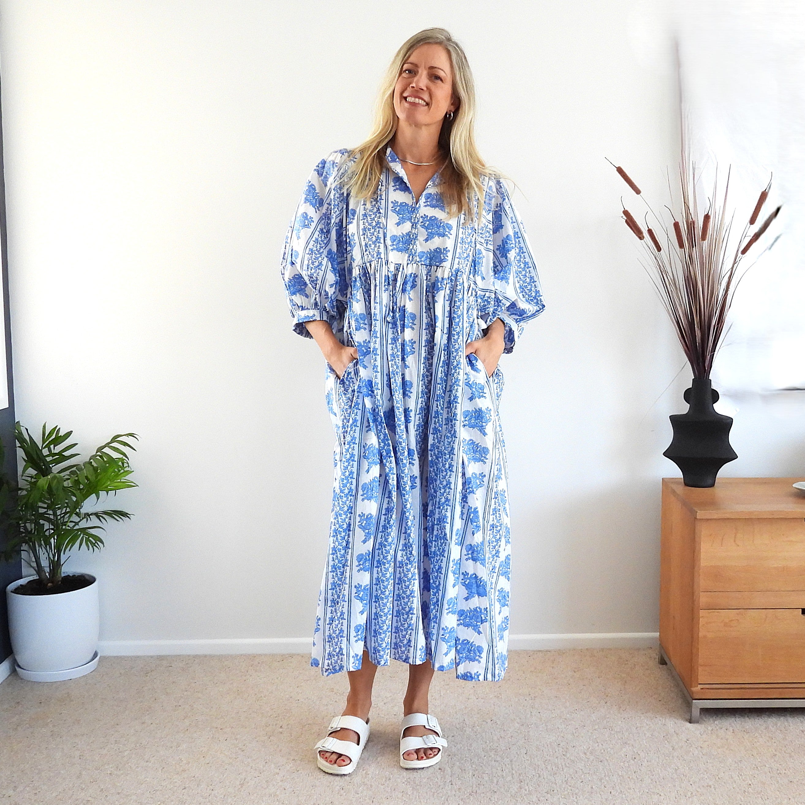 A woman stands indoors wearing a loose blue and white patterned midi dress with pockets and elbow-length sleeves. The room has a white wall, beige carpet, a potted plant on the left, and a wooden side table with a black vase on the right.