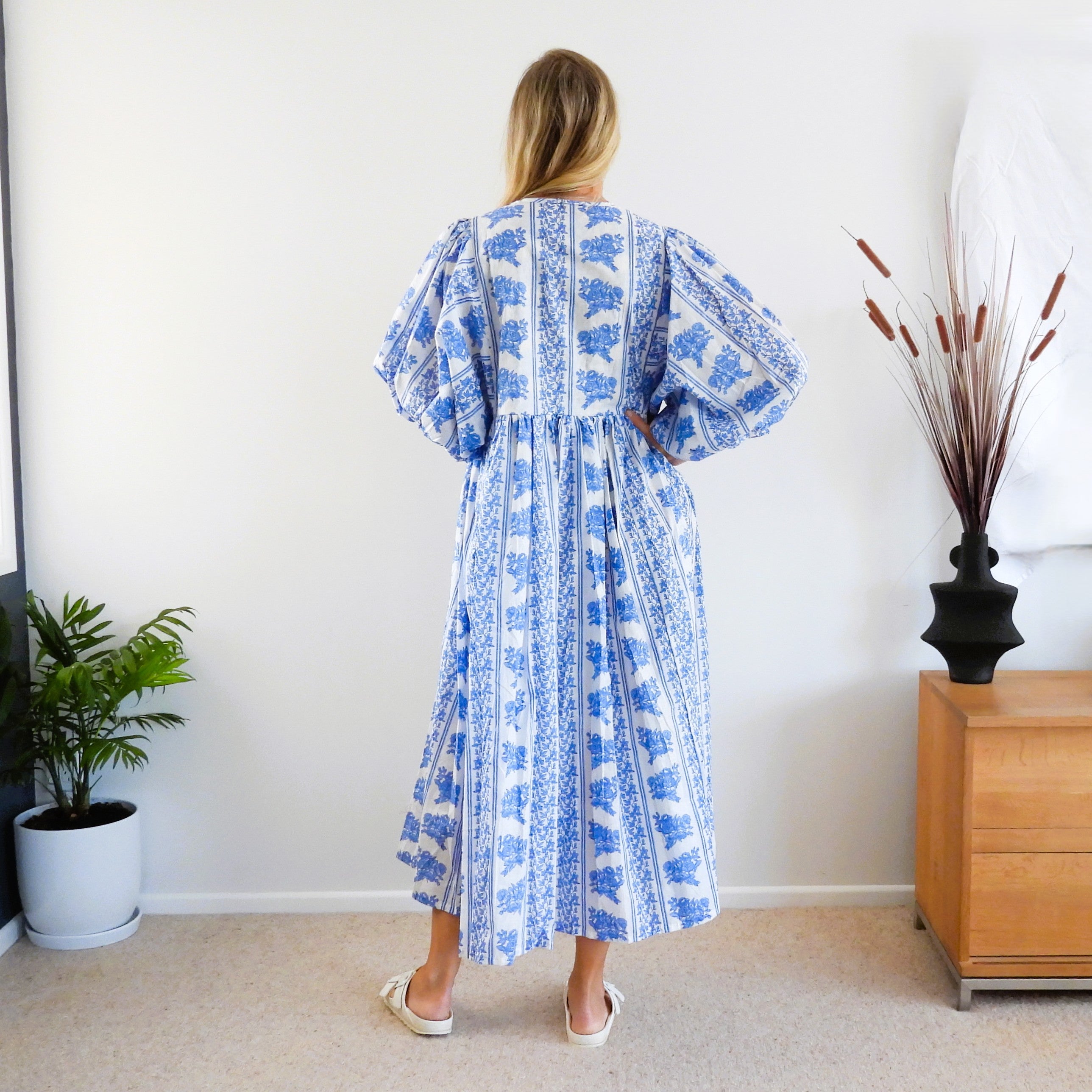 A woman stands with her back to the camera wearing a blue and white patterned midi dress with voluminous puff sleeves and a gathered waist.