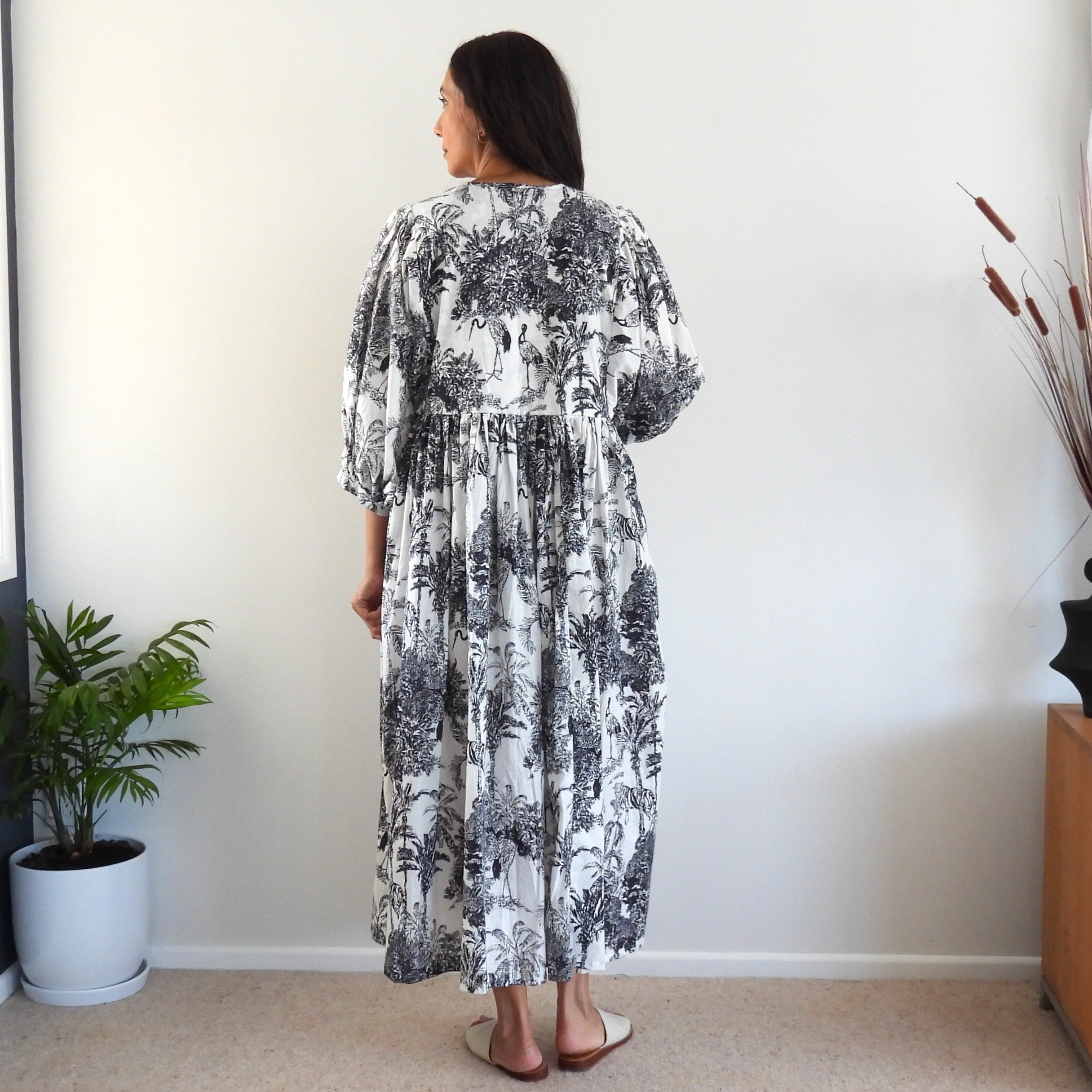 A woman stands with her back to the camera wearing a white midi dress with a black botanical toile pattern. The dress features three-quarter puffed sleeves and a gathered waist.