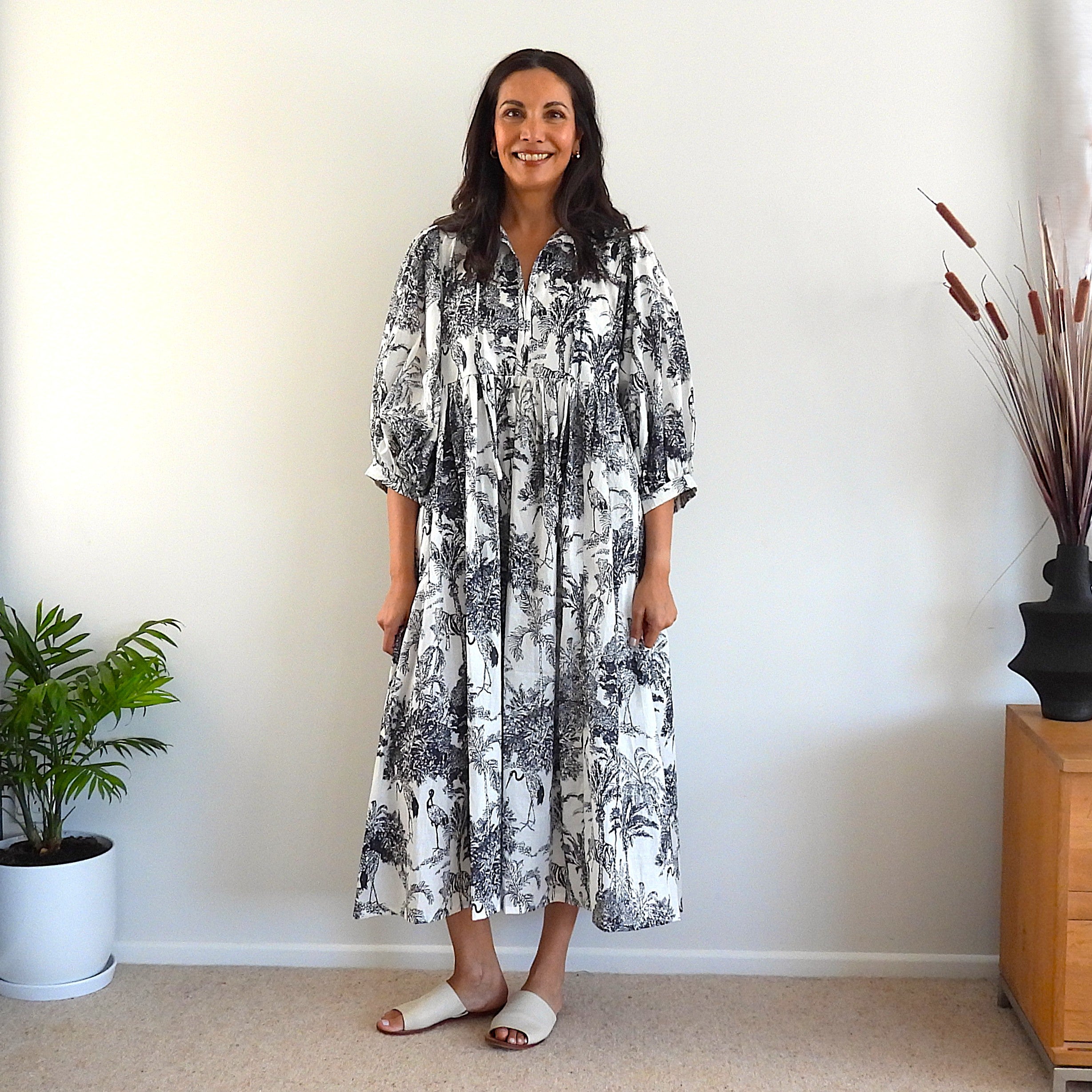 A woman stands indoors wearing a loose, knee-length dress with a black botanical and floral toile-style print on a white background. The dress has gathered sleeves and a gathered waist.
