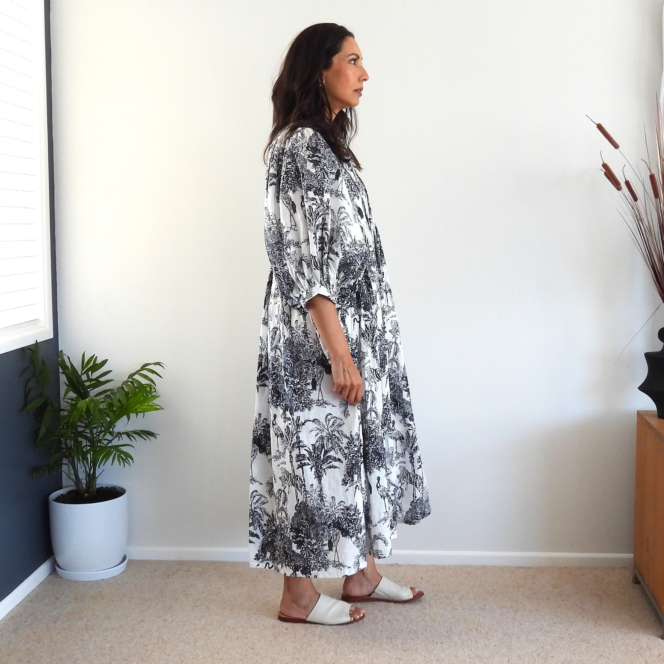 A woman stands in profile wearing a long-sleeve, knee-length white dress with a black botanical toile pattern. 