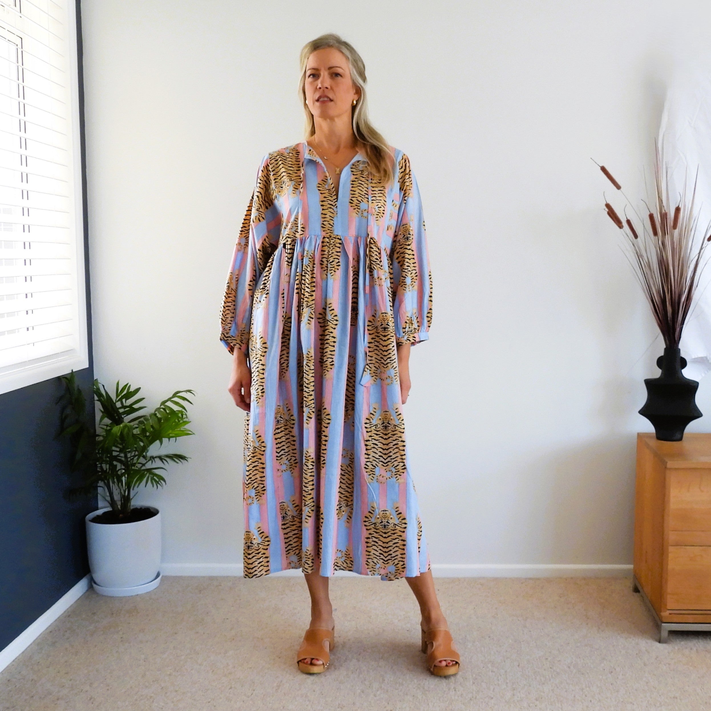 A woman stands indoors wearing a loose, long-sleeve midi dress with a light blue and pink wide stripes and brown abstract tiger print. The dress has a tie-up front and a relaxed fit.