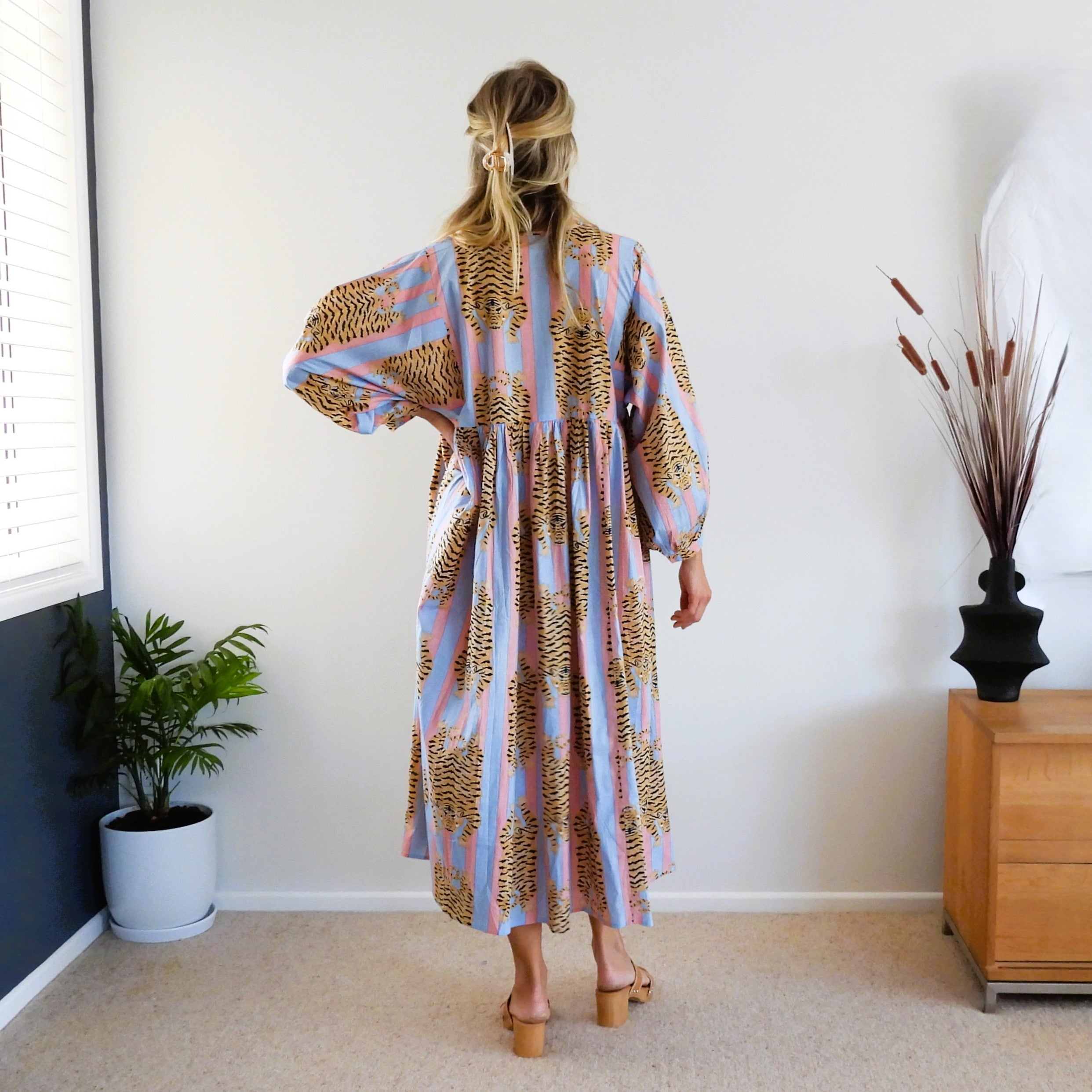 A woman viewed from behind wearing a long-sleeved maxi dress with a blue, pink and brown tiger pattern, standing in a brightly lit room with a window, potted plant, and wooden cabinet.