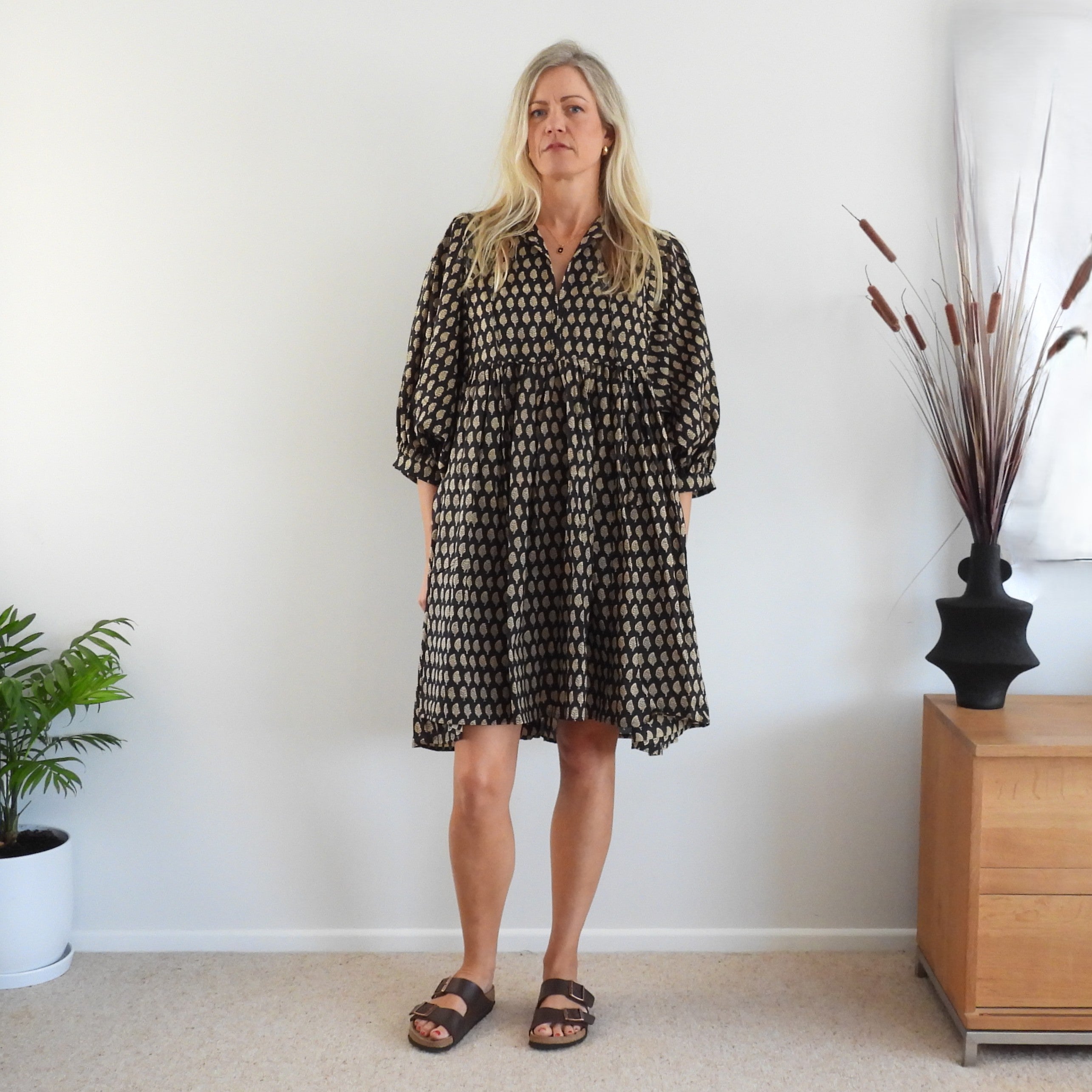 A woman stands in a room wearing a loose black dress with a beige geometric pattern, paired with brown slide sandals. 