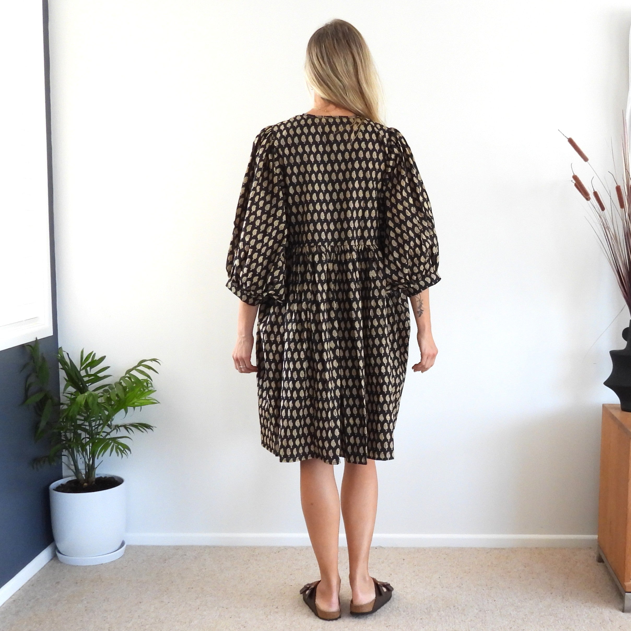 A woman stands with her back to the camera wearing a brown and black patterned dress with puff sleeves in a brightly lit room.