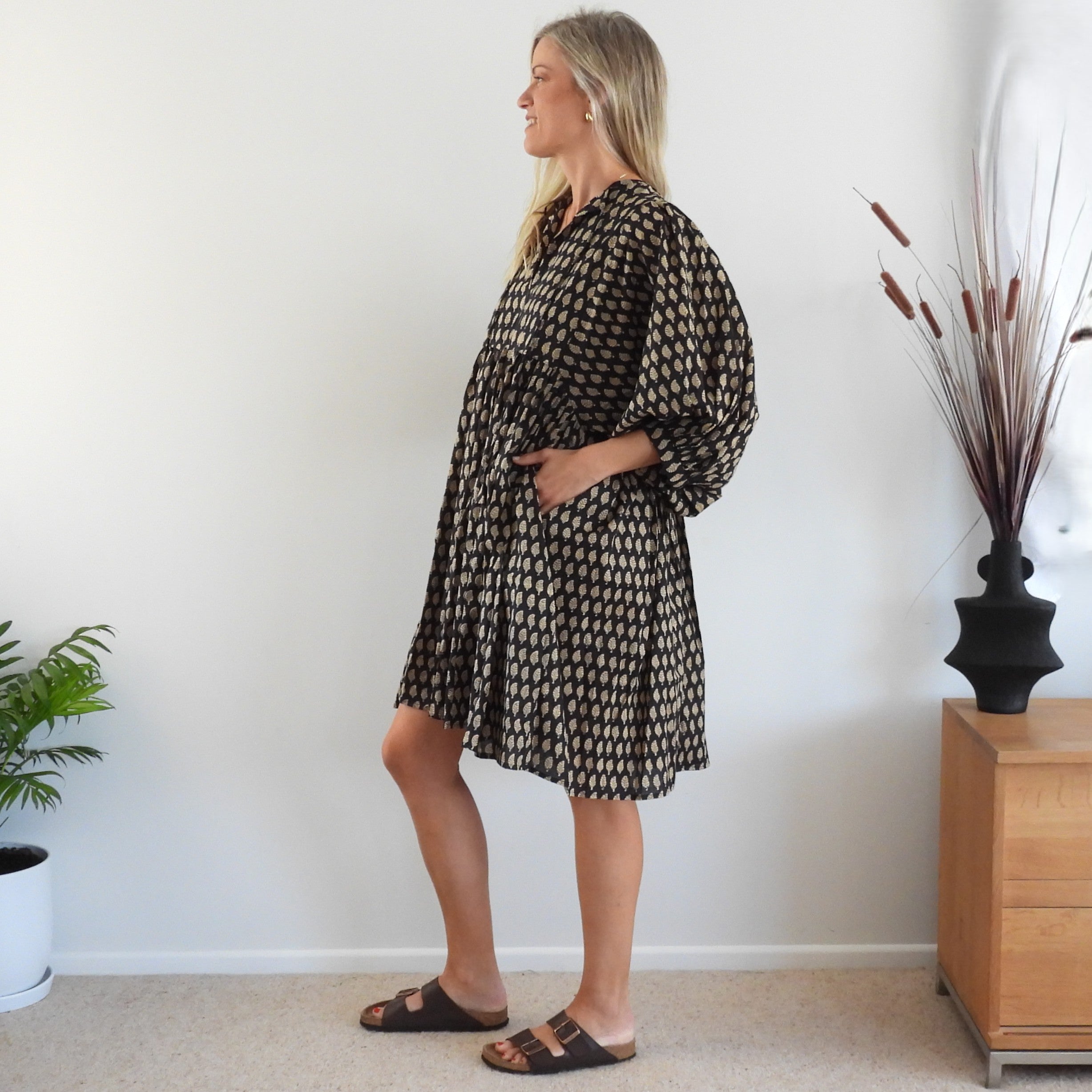 A woman modeling a loose-fitting black dress with a beige geometric pattern, standing in profile in a brightly lit room.